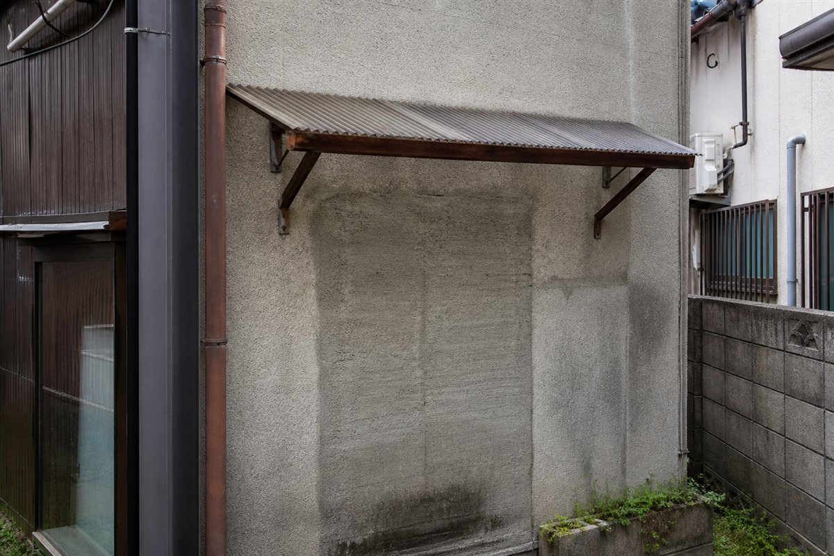 小さな波板の庇が、窓も扉もない補修済みの壁面だけを丁寧に覆っている