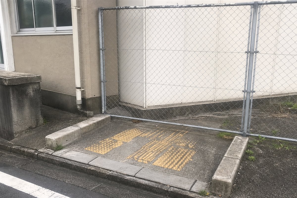 縁石の切り下げと短い車椅子用スロープが残るが、その正面を新しい金網フェンスが完全に塞いでいる